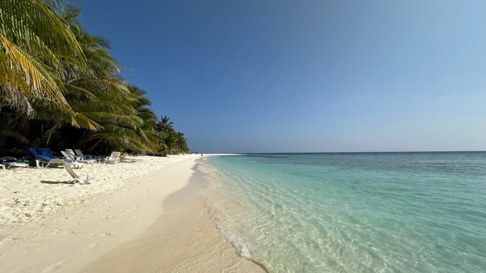 weisser Strand mit Palmen und türkis blauem seichten Meerwasser, mit ein paar Sonnenliegen am Strand, menschenleer, blauer Himmel